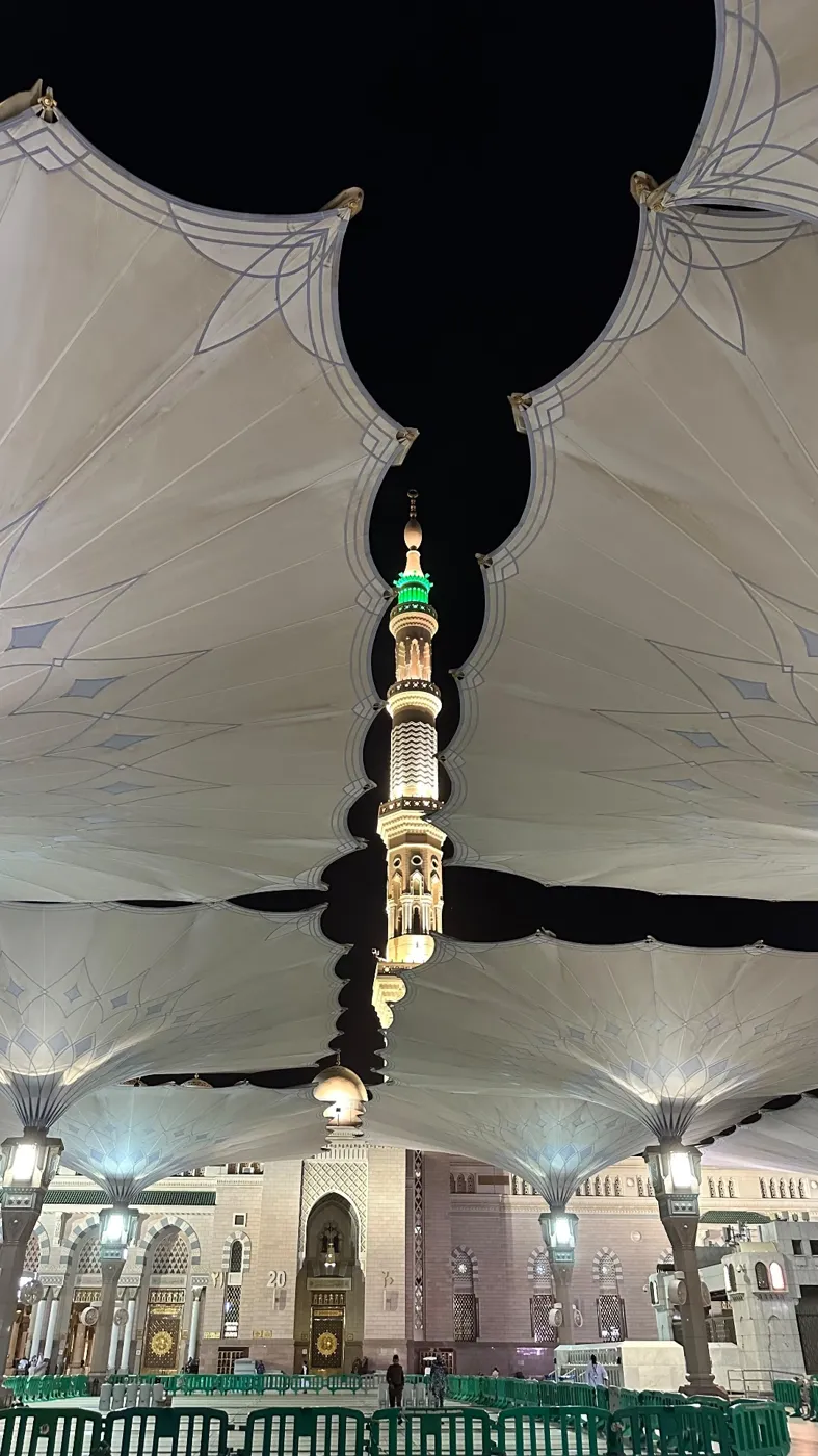 An illuminated minaret of Masjid an-Nabawi seen through the open giant umbrella canopies at night