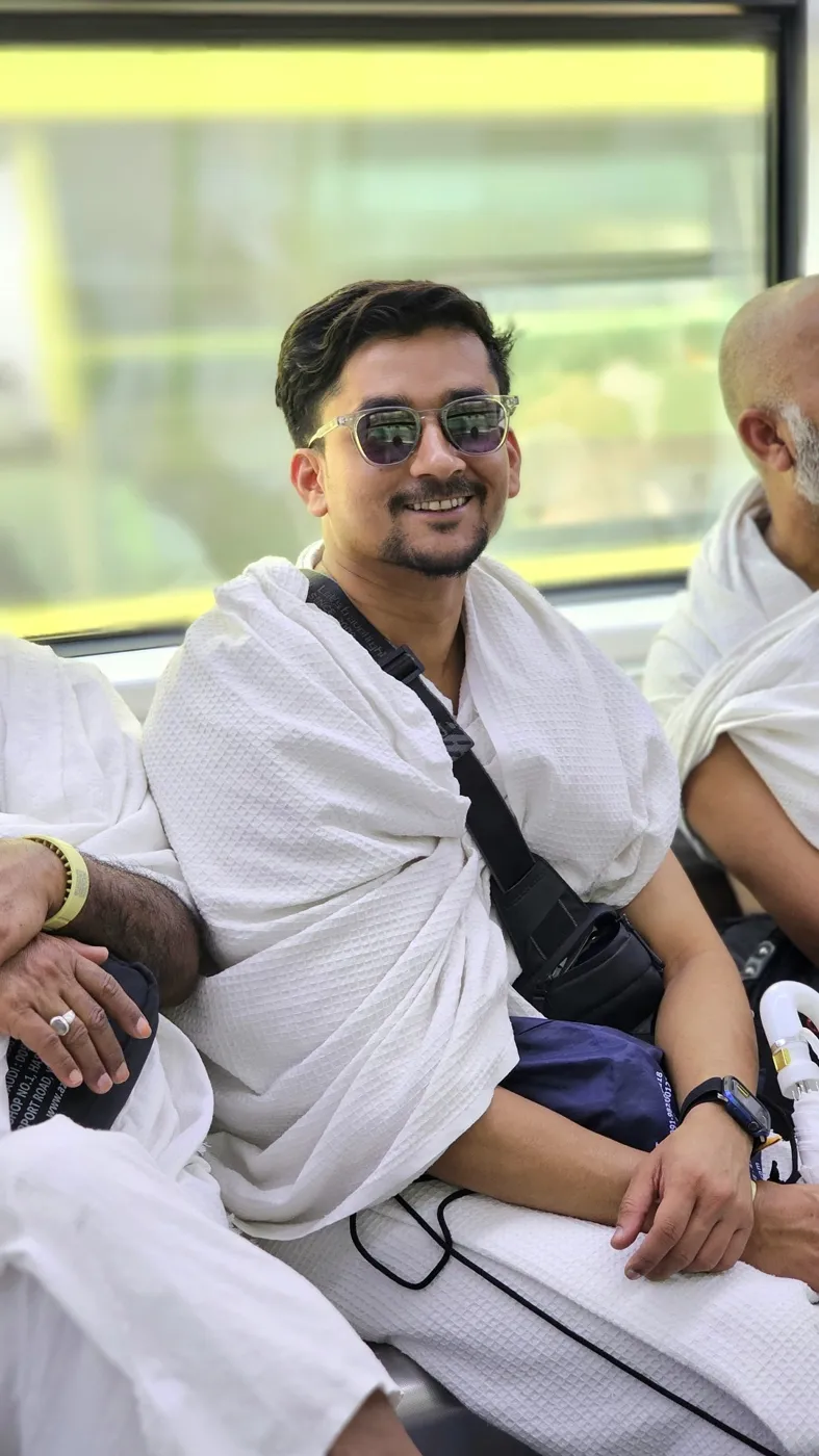 A smiling young pilgrim in white Ihram seated among fellow pilgrims on a train