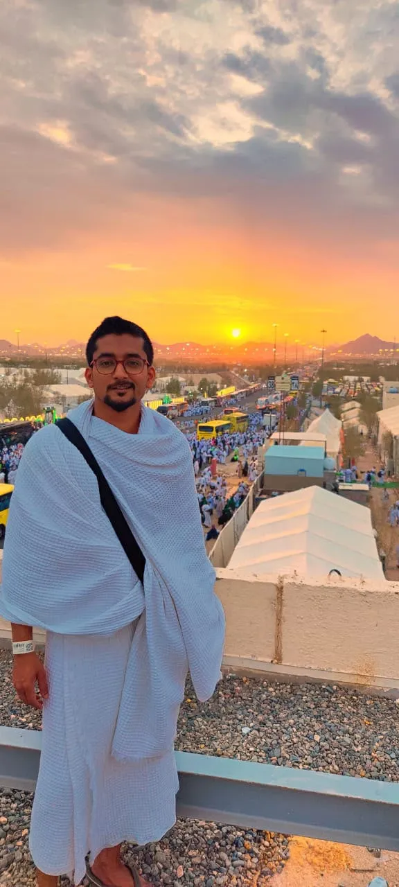 A pilgrim in white Ihram standing on a rooftop at sunset, with crowds of pilgrims, buses and tents below